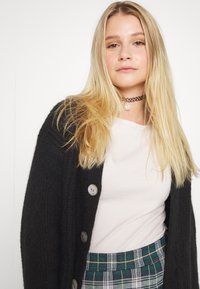 Černý pletený kardigan s velkými knoflíky přes světlý top, doplněný zelenou a modrou kostkovanou sukní. Dlouhé blond vlasy rámují obličej.