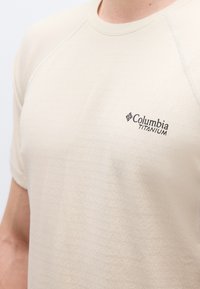 Κοντινό πλάνο ενός ατόμου που φοράει μπεζ κοντομάνικο πουκάμισο Columbia Titanium με διακριτό υφασμάτινο μοτίβο και ορατές ραφές στα μανίκια.