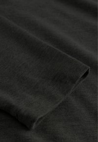 Tumši zaļš kokvilnas audums ar gludu tekstūru, ar salocītu piedurkņu malu un redzamām šuvēm. Tuvplāns uz auduma.