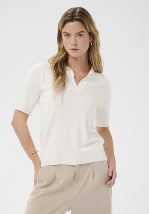 Ženska z dolgimi blond lasmi, oblečena v belo polo majico s kratkimi rokavi in bež hlače, stoji z eno roko v žepu pred belim ozadjem.