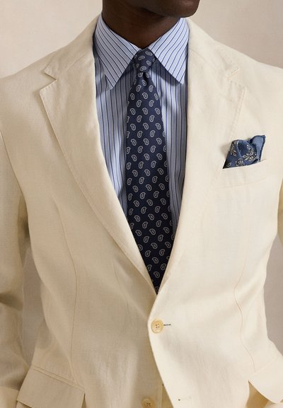 Lininis, kreminis švarkas ant šviesiai mėlynos dryžuotos marškinių. Tamsiai mėlynas kaklaraištis su paislio raštu ir mėlynas kišenės kvadratas su gėlių akcentais.