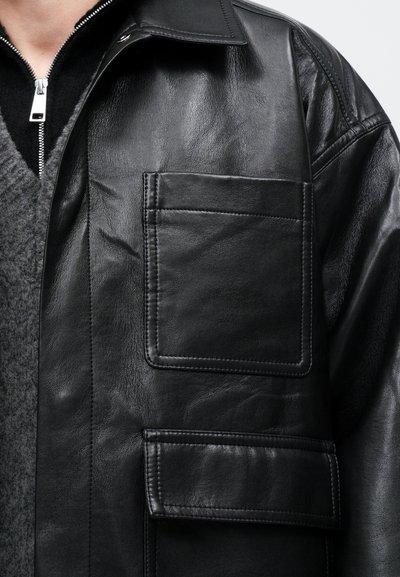 Primo piano di una giacca di pelle nera con due tasche cucite indossata sopra un maglione di lana grigio e un indumento con zip nera.