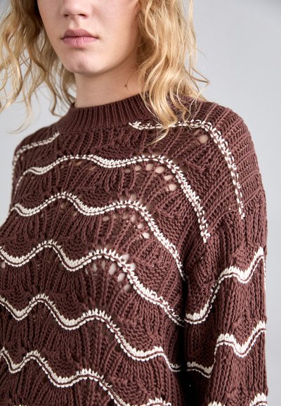 Brun strikket sweater med et bølgemønster og cremefarvede accenter. Designet inkluderer gennemsigtige sektioner og en ribbet halsudskæring.