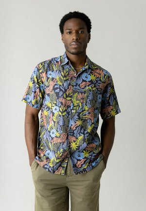 Mężczyzna ubrany w kolorową, krótkorękawową koszulę na guziki z kwiatowymi i zwierzęcymi wzorami, stojący z rękami w kieszeniach na jednolitym tle.