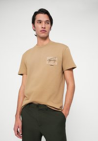 Mees, kellel on seljas helepruun Tommy Jeans'i T-särk ja tumerohelised püksid, seisab ühel käel taskus, vaadates kõrvale ühtlasel taustal.