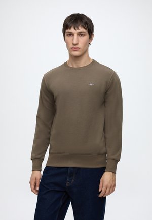 Jeune homme aux cheveux bruns courts portant un sweat-shirt marron uni à manches longues et un jean bleu foncé, debout devant un fond uni.