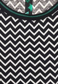 Černobílá látka se zigzag vzorem, s pevným černým výstřihem a zelenými hvězdnými akcenty. Měkká textura, vhodná na volnočasové nošení.