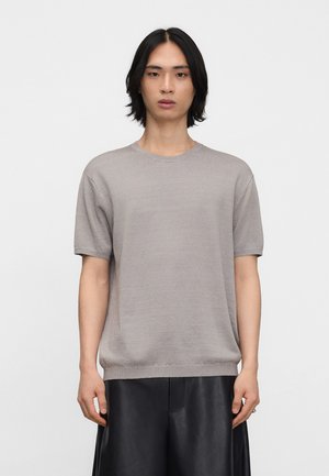 Giovane con capelli neri di media lunghezza che indossa una maglia grigia a maniche corte e pantaloni neri a gamba larga, in piedi davanti a uno sfondo bianco.