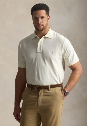 Rövid göndör hajú, szakállas férfi, fehér galléros inget visel, amelyet barnás nadrágba tűrt be, barna övvel és karórával.