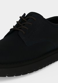 Chaussure noire à lacets avec une semelle épaisse et texturée et une finition lisse et mate visible sur les parties latérales et avant.