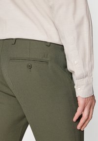 Olivgrön skräddarsydda byxor med ett subtilt herringbone-mönster, med en ficka med lock och knappstängning samt en beige skjorta med knappar.