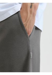 Grå sweatpants med en glat tekstur, der har en sidelomme, synlig stikning og et lille mærkeskilt på sømmen.