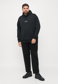 Czarna bluza z kapturem z kieszenią kangurzą, z logo na klatce piersiowej. W zestawie z czarnymi spodniami i czarnymi sneakersami. Gładka tekstura materiału.