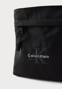 Borsa nera in nylon con chiusura a zip, dotata di tracolla intrecciata e logo "Calvin Klein" ricamato in argento. Texture liscia e forma strutturata.
