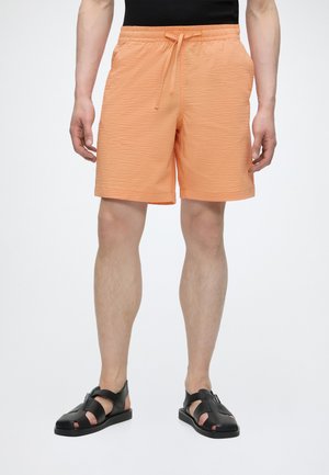 Homme portant un short orange clair texturé à cordon de serrage et des sandales en cuir noir, debout devant un fond blanc uni.