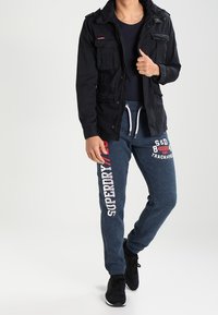 Zwarte utilityjas met vier zakken, gecombineerd met marineblauwe sweatpants met witte letters en een trekkoord in de taille. Zwarte sneakers maken de outfit compleet.