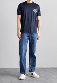 Námořnický bavlněný tričko s bílým logem "Emporio Armani 1981", v kombinaci se světle modrými džíny a bílými teniskami s modrými akcenty.