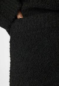 Tissu noir texturé avec une surface duveteuse, comportant une poche avec une main visible qui dépasse. Accent sur la douceur et le détail du matériau.