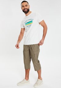 Witte katoenen t-shirt met blauwe en gele print met de tekst 'SAINT TROPEZ'. Olijfgroene cargo shorts met zakken en een cropped ontwerp. Beigekleurige sneakers.
