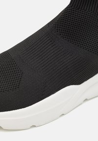Černé pletené tenisky s texturovaným svrškem, které mají design ponožky a bílou gumovou podrážku. Minimalistická a bezešvá konstrukce.