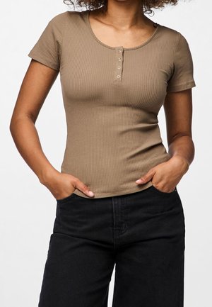 Femme portant un haut henley beige moulant à manches courtes côtelées avec des boutons pression, un pantalon noir, les mains dans les poches, sur un fond uni.
