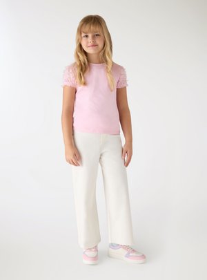Młoda dziewczyna stoi, ubrana w różowy top z krótkim rękawem z fakturowanymi rękawami, białe szerokie spodnie i pastelowe tenisówki.