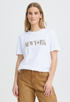 Jeune femme aux cheveux blonds et raides portant un T-shirt blanc imprimé "NEW YORK" et un pantalon marron, debout devant un fond clair uni.