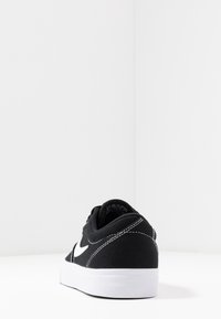 Baskets noires à tige basse avec semelle blanche et swoosh Nike blanc, montrées de l'arrière sur une surface blanche avec un fond blanc uni.