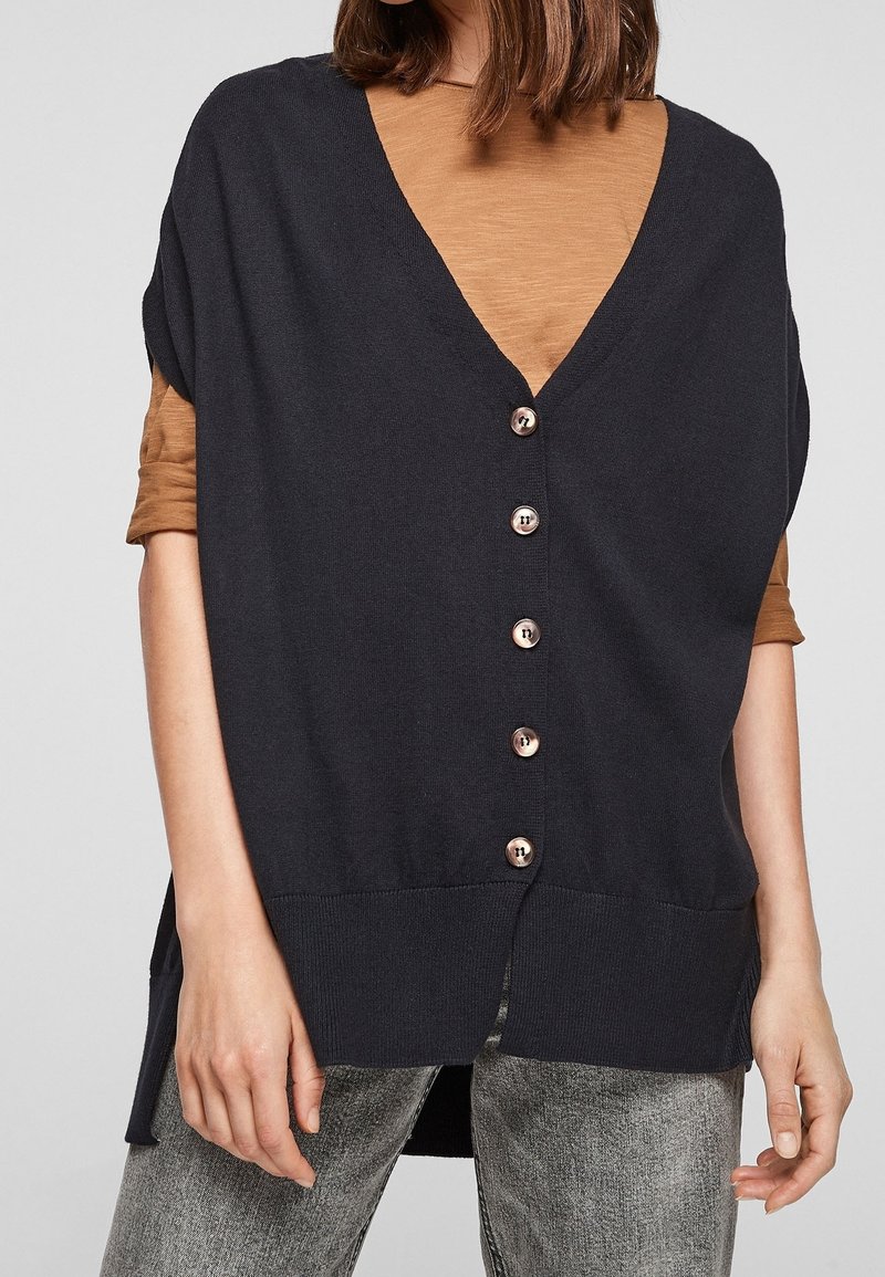 Mörk marinblå cardigan med djup V-halsringning och korta ärmar, prydd med sex metallknappar fram. Bärs över en brun topp.