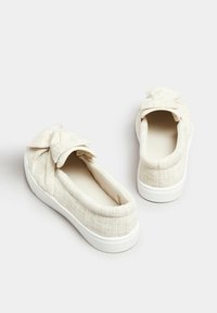 Scarpe slip-on in tela beige con tessuto texture, dotate di un dettaglio a fiocco sulla parte anteriore e una suola in gomma bianca.