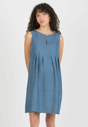 Kobieta o kręconych włosach w niebieskiej plisowanej sukience bez rękawów z małym rozcięciem przy dekolcie na jednolitym jasnym tle.