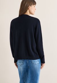 Femme aux longs cheveux bruns portant un pull noir uni et un jean bleu clair, vue de dos contre un mur beige clair.