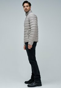 Chaqueta acolchada gris claro con cuello alto y costuras horizontales visibles, combinada con jeans negros y zapatos negros.