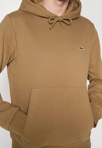 Brun hoodie i mjukt tyg med kenguruficka, dragsko i huvan och en liten broderad logotyp på bröstet.