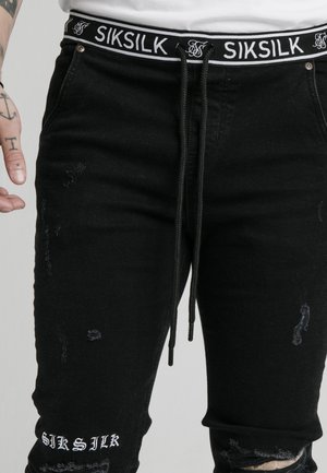 Schwarze abgenutzte Jeans mit Siksilk-Logo am Bund und nahe am Knie, getragen von einer Person mit Tätowierungen an linker Hand und Handgelenk.