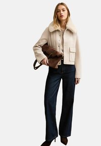 Kobieta w kremowej kurtce z kołnierzem z baranka, beżowym swetrze, ciemnych dżinsach o szerokich nogawkach, brązowych spiczastych botkach, trzymająca dużą brązową zamszową torebkę.