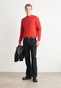 Punane villane sviiter ümmarguse kaelusega, mustad teksad ja mustad nahkkingad. Modell hoiab musta nahkjahtumist. Minimalistlik taust.