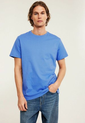 Junger Mann steht, trägt ein schlichtes blaues T-Shirt und blaue Jeans, mit einer Hand in der Tasche vor einem neutralen Hintergrund.