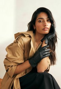 Guanti in pelle nera con tre cuciture e un design elegante, abbinati a un trench beige e un abito scuro. Texture liscia e forma aderente.