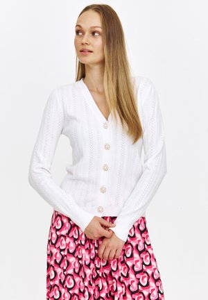 Biała dziergana kamizelka z dekoltem w kształcie litery V, z zapięciem na guziki i teksturowanym wzorem, w połączeniu z różową spódnicą w wzory.