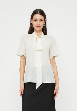 Mujer con pelo largo y oscuro lleva una blusa blanca de manga corta con un lazo delantero y una falda negra, de pie frente a un fondo liso.