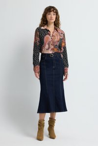 Larga falda vaquera azul con una abertura frontal y trabillas para cinturón, combinada con una camisa de manga larga estampada en tonos de rosa y negro. Botines marrones hasta el tobillo.