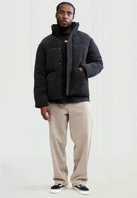 Homem vestido com uma jaqueta acolchoada preta, camisa preta, calças bege e ténis pretos, em pé com uma mão no bolso da jaqueta, contra um fundo simples.