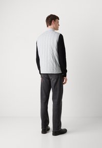 Colete acolchoado cinza claro com um colarinho alto, usado sobre uma camisa preta de mangas compridas. Combinado com jeans cinza escuro e sapatos pretos.