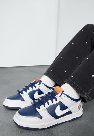 Person iført hvide og marineblå Nike-sneakers med orange logo, hvide sokker og sorte bukser med små sølvnitter, siddende på gråt gulv.