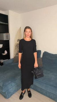 Melna pieguļoša kleita ar trīs ceturtdaļu piedurknēm un sānu sprādzes detaļu, kopā ar melnām loafers kurpēm un strukturētu rokassomu. Teksturēts zilas dīvāna fons.