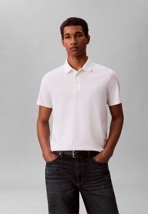 Młody mężczyzna ubrany w białą koszulkę polo z krótkim rękawem wpuszczoną w ciemne jeansy, ręce w kieszeniach na jednolitym szarym tle.