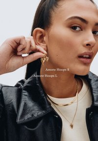 Goldene Ohrhänger in Herzform, getragen von einem Model mit langen Haaren. Der Text lautet "Amore Hoops S" und "Amore Hoops L." Das Model trägt eine schwarze Jacke und einen cremefarbenen Pullover.