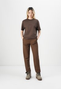Tricou scurt cu mânecă închisă la culoare, cu logo, asociat cu pantaloni maro. Modelul poartă bocanci bej cu șireturi; fundalul simplu evidențiază ținuta.
