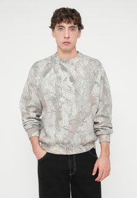 Γκρι φούτερ με καμουφλάζ σχέδιο από κλαδιά και φύλλα, με άνετη εφαρμογή και ριμπ μανσέτες. Φοριέται με μαύρο παντελόνι.